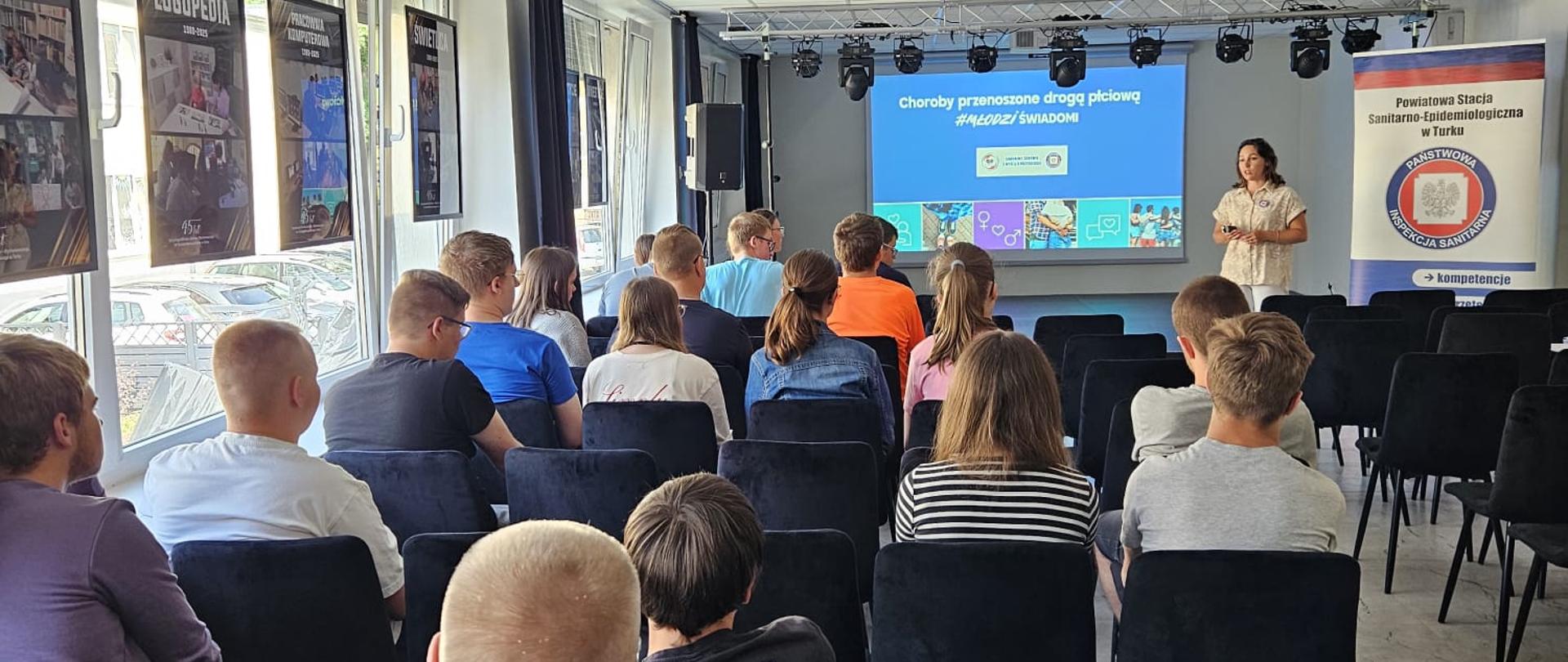 młodzież siedząca na krzesłach w sali konferencyjnej, przed nimi kobieta prowadząca wykład