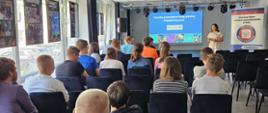 młodzież siedząca na krzesłach w sali konferencyjnej, przed nimi kobieta prowadząca wykład