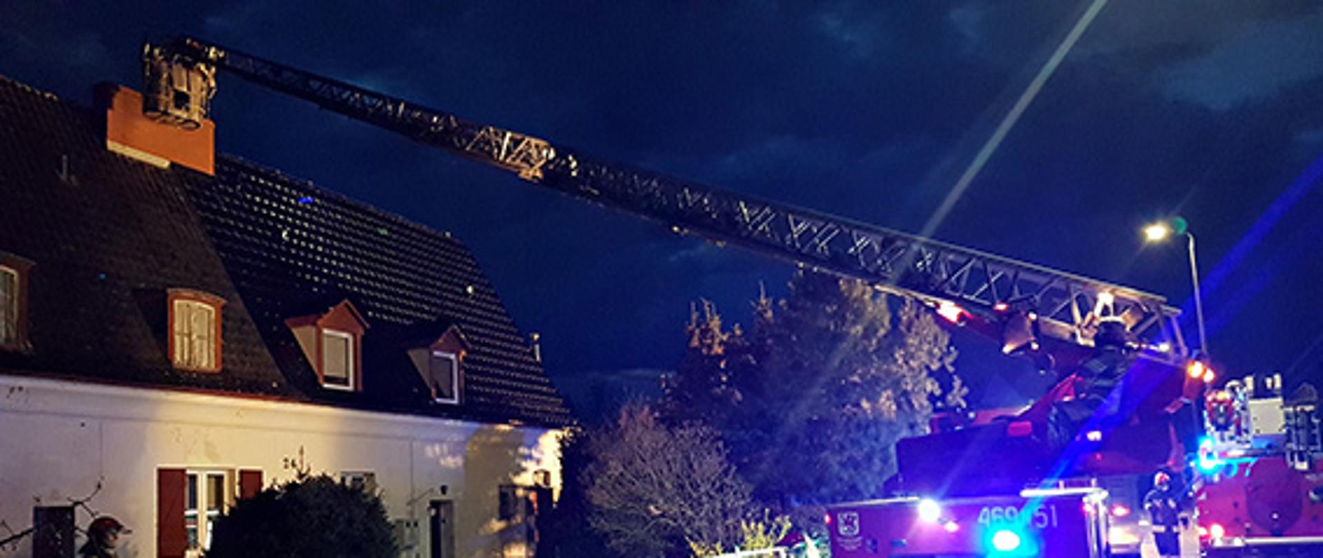 Na zdjęciu widać działania strażaków w porze nocnej. Dwóch strażaków przy pomocy strażackiej drabiny gasi pożar sadzy w kominie.
