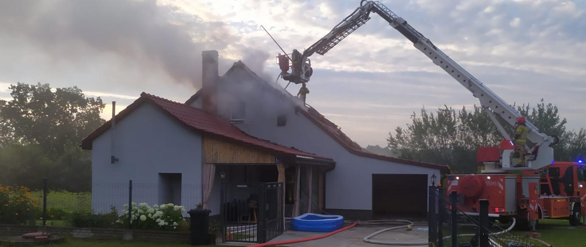 Zdjęcie przedstawia pożar budynku jednorodzinnego w Rąbinie, przy budynku znajduje się drabina strażacka z której strażak podaje prąd wody. Po podwórku rozwinięte są linie gaśnicze.