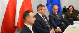Przy stoliku siedzi trzech mężczyzn w garniturach i ubrana na czarno kobieta, za nim dwie polskie flagi i jedna flaga UE, dalej białe ściany.