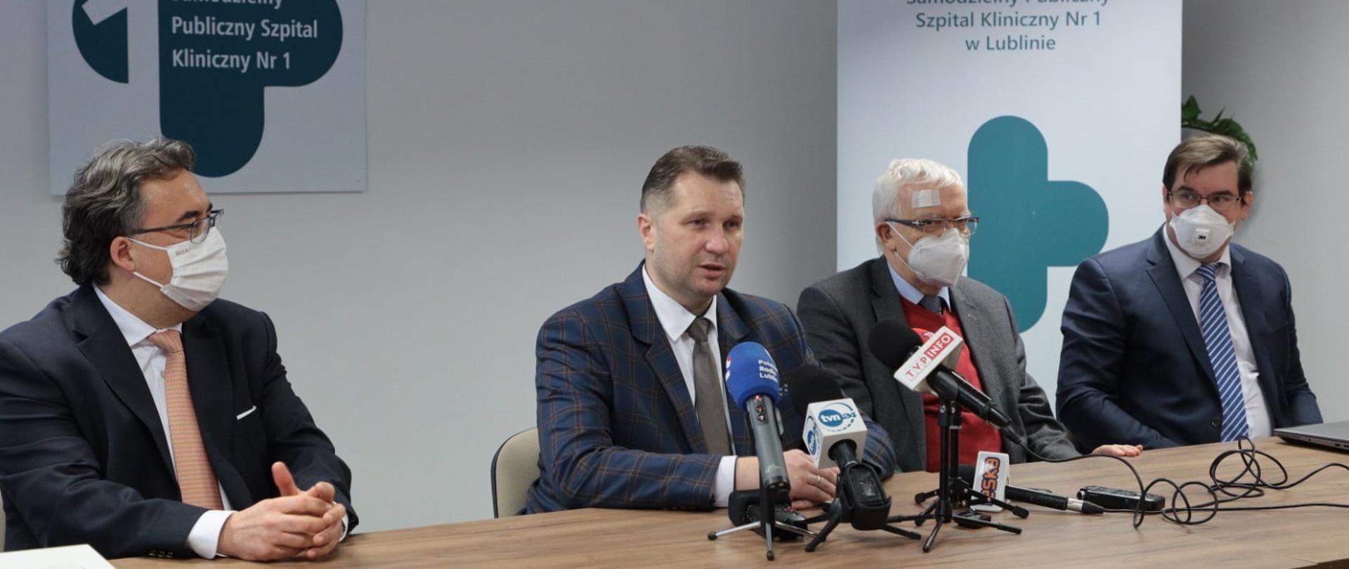 Przy stole siedzi kilku mężczyzn ubranych w garnitury. Na twarzach mają maseczki. Przed ministrem Czarnkiem ustawionych na stole jest kilka mikrofonów. W tle banery Samodzielnego Publicznego Szpitala Klinicznego nr 1 w Lublinie. 