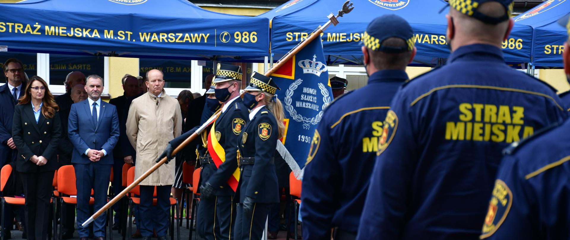 Na pierwszym planie zdjęcia znajdują się pododdziały uformowane z funkcjonariuszy Straży Miejskiej Miasta Stołecznego Warszawy. Na drugim planie, tuż przed trybuną honorową przechodzi poczet sztandarowy Straży Miejskiej Miasta Stołecznego Warszawy. 