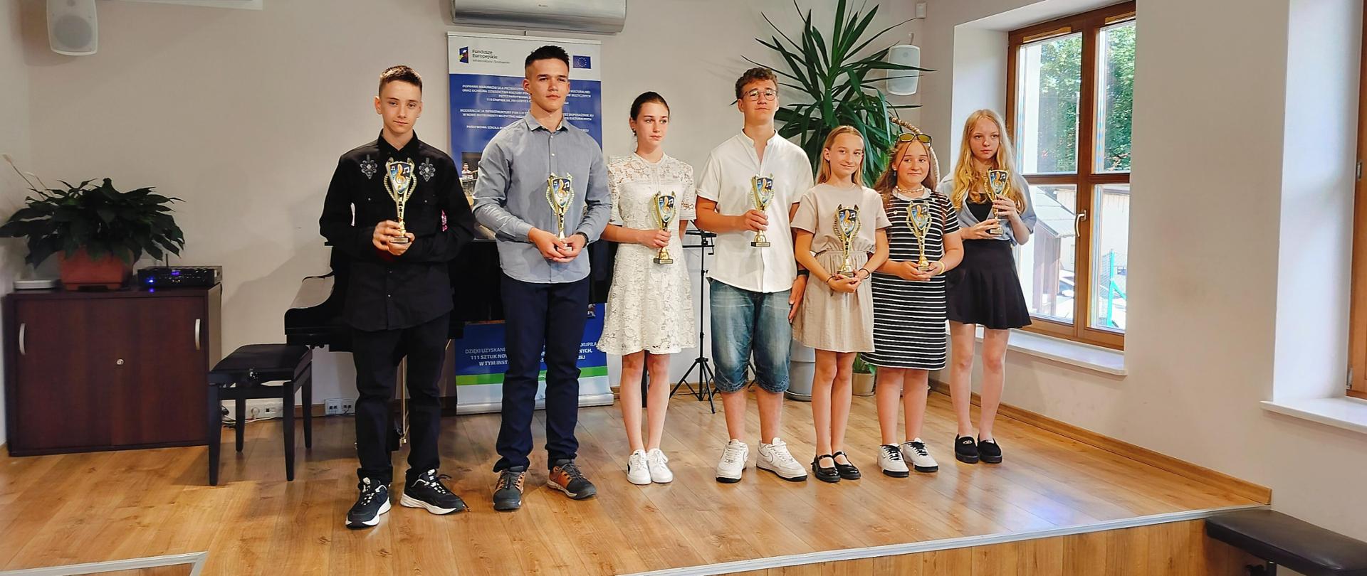 Sala kameralna, siedmioro dzieci, trzech chłopców oraz czwórka dziewczyn stoi na podium z pucharami. Za nimi od lewej strony: brązowa szafka, krzesło do fortepianu oraz fortepian, baner reklamowy pulpit oraz kwiatek. Pod sceną po prawej stronie krzesło do fortepianu.