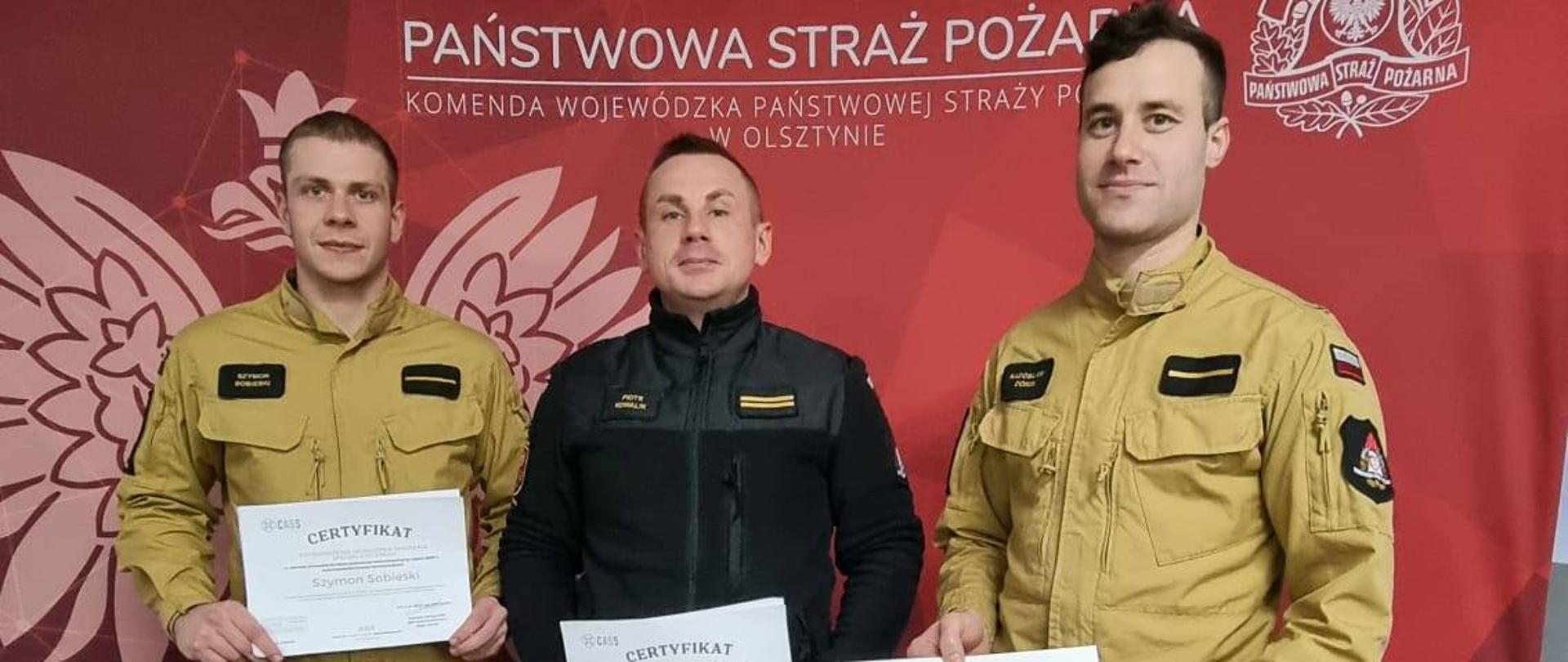 Trzech strażaków na tle czerwonej ścinaki z napisem państwowa straż pożarna. Strażacy trzymaj w rekach dyplomy. Pozują do zdjęcia.