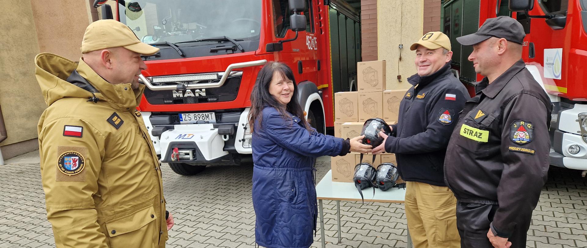 Na zdjęciu widać moment przekazania sprzętu strażackiego przed remizą. Kobieta w granatowym płaszczu wręcza strażakowi maskę ochronną Dräger FPS 7000. Obok nich stoją dwaj inni strażacy w mundurach — jeden w czarnym, drugi w beżowym. W tle znajdują się dwa czerwone wozy strażackie oraz stół, na którym ułożono kilka masek i kartonowych pudełek z tym samym sprzętem. Wszyscy uczestnicy wyglądają na zadowolonych.