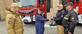 Na zdjęciu widać moment przekazania sprzętu strażackiego przed remizą. Kobieta w granatowym płaszczu wręcza strażakowi maskę ochronną Dräger FPS 7000. Obok nich stoją dwaj inni strażacy w mundurach — jeden w czarnym, drugi w beżowym. W tle znajdują się dwa czerwone wozy strażackie oraz stół, na którym ułożono kilka masek i kartonowych pudełek z tym samym sprzętem. Wszyscy uczestnicy wyglądają na zadowolonych.