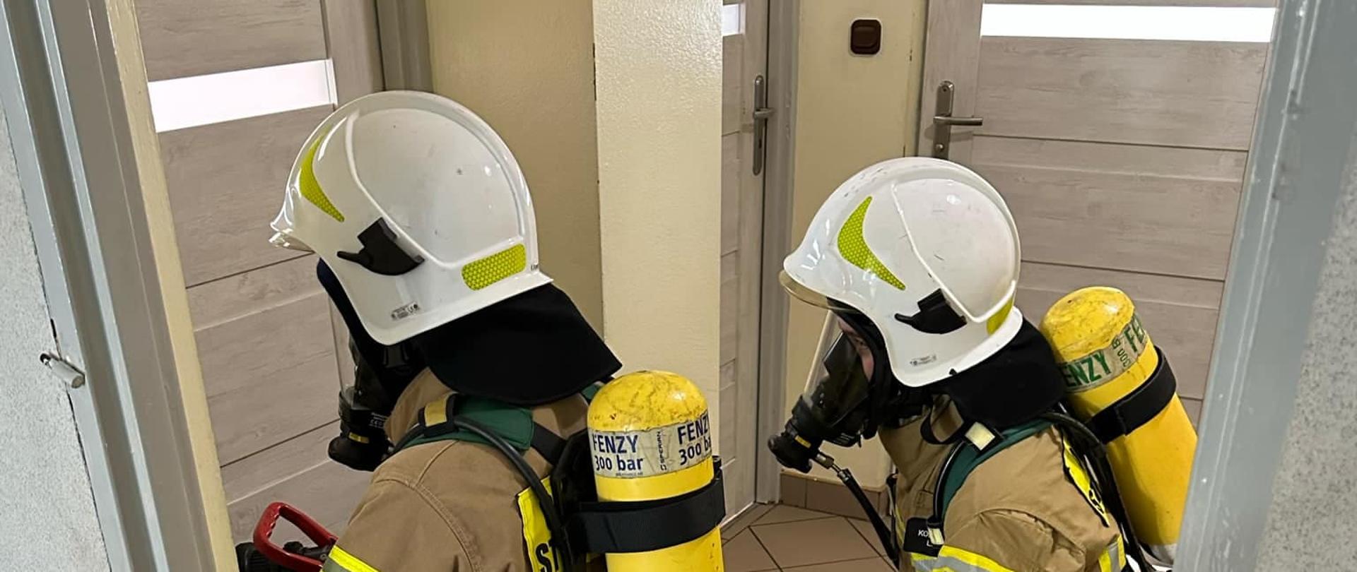 Strażacy w ubraniach specjalnych i hełmach klękają przed drzwiami, 