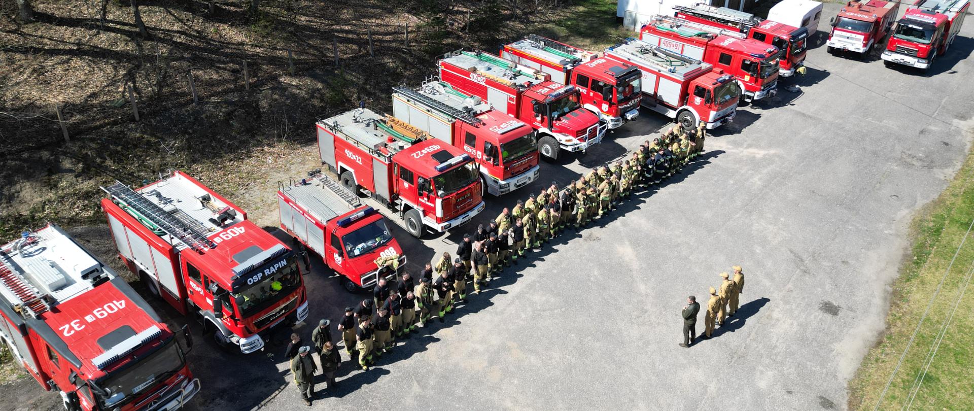 Odprawa po zakończonych ćwiczeniach. Na zdjęciu strażacy oraz pojazdy ratowniczo- gaśnicze.