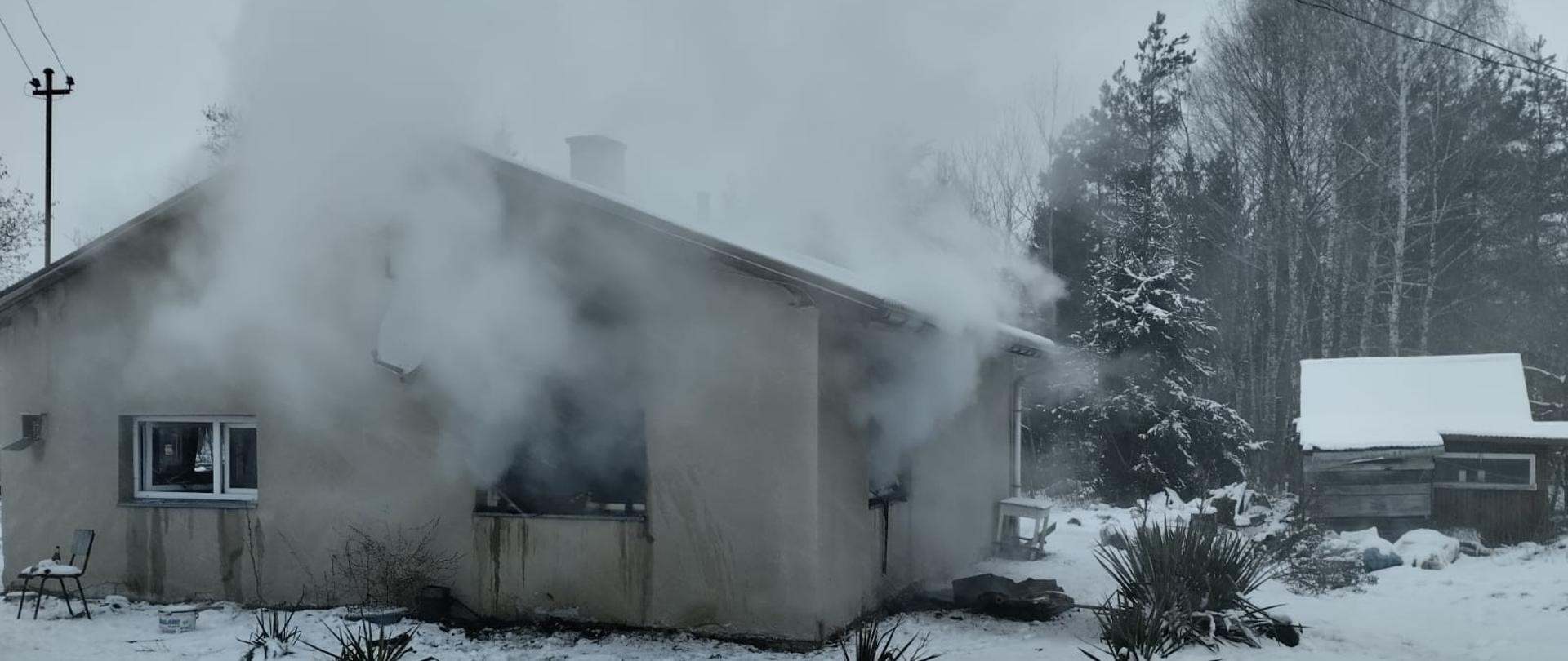 Dom z którego wydobywa się dym, na pierwszym planie widać wąż strażacki zakończony prądownicą.