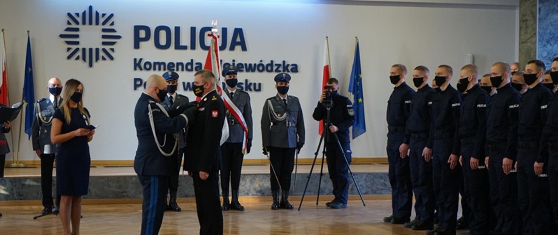 Komendant wojewódzki Policji przypina medal pomorskiemu komendantowi wojewódzkiemu Państwowej Straży Pożarnej podczas ceremonii ślubowania nowo przyjętych policjantów. W tle stoją policjanci.