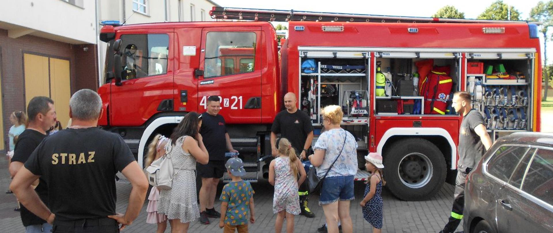 Na zdjęciu dorośli oraz dzieci słuchające wykładu strażaka. W tle pojazd pożarniczy z otwartymi skrytkami.