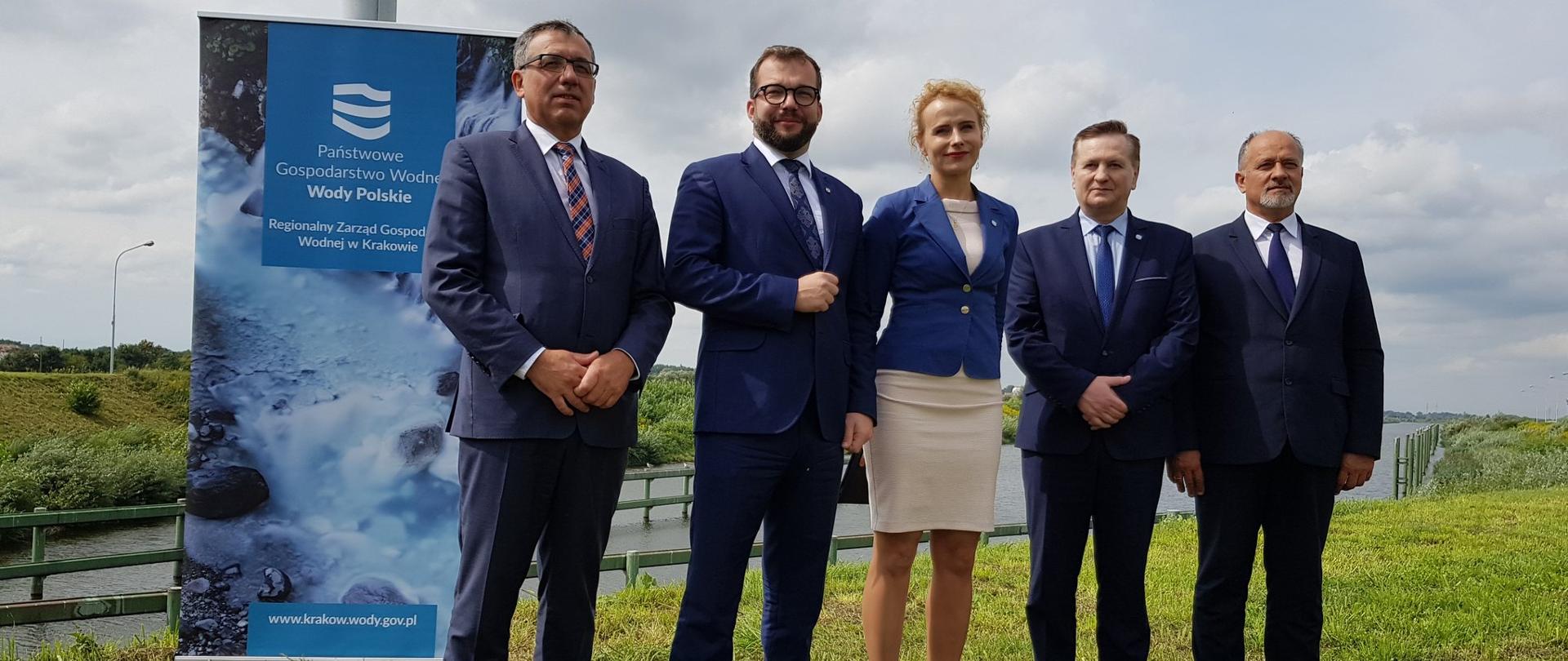 Minister Puda stoi z czterema pozostałymi uczestnikami spotkania, w tle płynie rzeka, za prelegentami stoi ścianka promująca "Wody Polskie". 