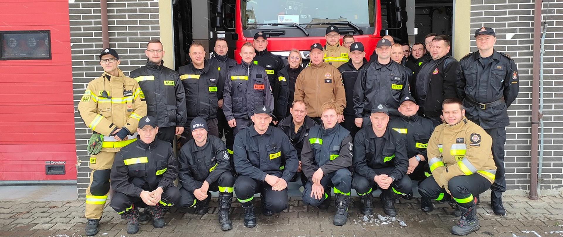 Dzień Widok na otwartą bramę garażową. W bramie stoi samochód ratowniczo - gaśniczy w kolorze czerwonym. Przed samochodem, w otwartej bramie stoi kilkanaście osób ubranych w ubrania koszarowe w kolorze czarnym na których naszyte są elementy odblaskowe w kolorze żółtym. Kilka osób ubrana w ubranie specjalne w kolorze piaskowym. 