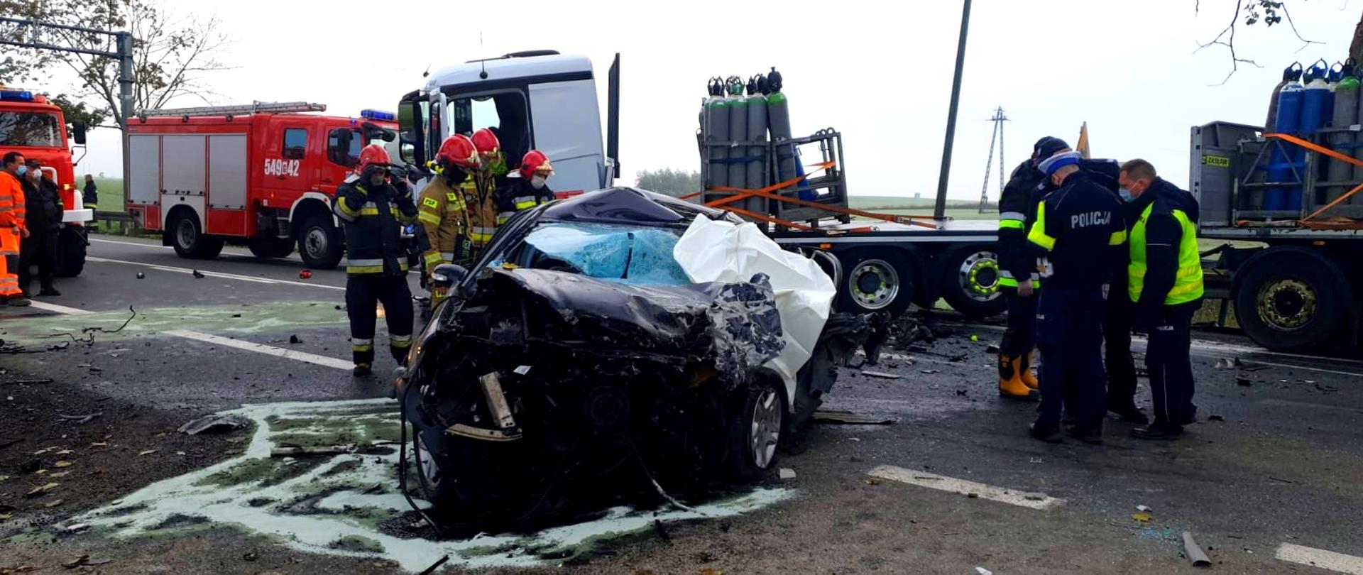 Zdjęcie wypadku komunikacyjnego z udziałem samochodu ciężarowego przewożącego butle z gazami technicznymi oraz samochodu osobowego. Na zdjęciu widoczne wraki pojazdów oraz osoby ze służb prowadzące działania na miejscu.