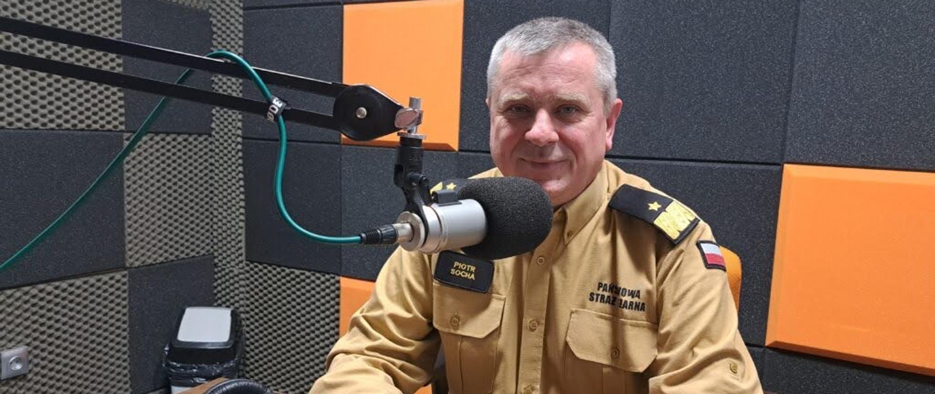 Funkcjonariusz Państwowej Straży Pożarnej siedzi przed mikrofonem w studiu przed nil leżą słuchawki zanim znajduje się kosz.