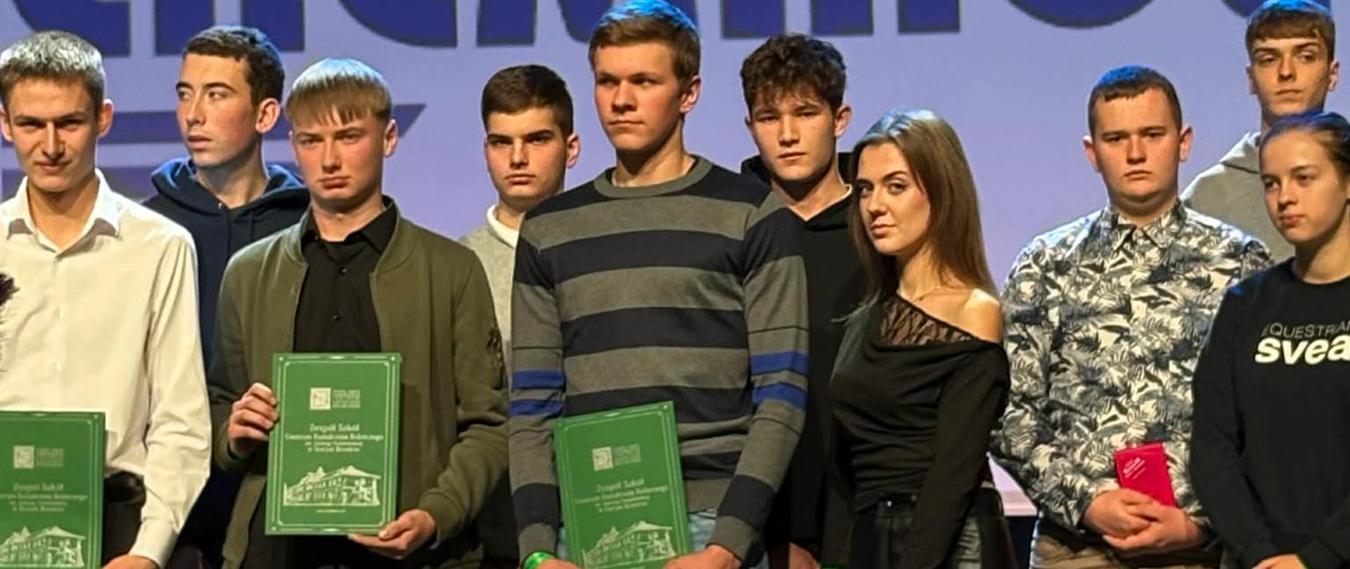 Grupa młodych ludzi bierze udział w ceremonii wręczenia nagród. Na tle widocznego logotypu firmy Chemirol, trzymają dokumenty i torby z logo sponsorów wydarzenia