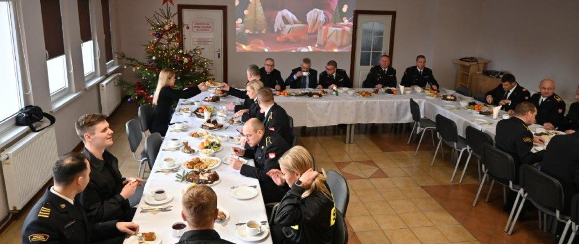 1. Funkcjonariusze w mundurach i pracownicy cywilni siedzą przy stołach. Na stołach są białe obrusy i jedzenie. Na środku na ścianie jest wyświetlony obraz z rzutnika. Na obrazie widać prezenty. Po lewej stronie w rogu stoi choinka. Obok choinki po lewej stronie są drzwi.
Po prawej stronie są drzwi i mównica. Po lewej stronie są okna. Ściany pomieszczenia są białe.
