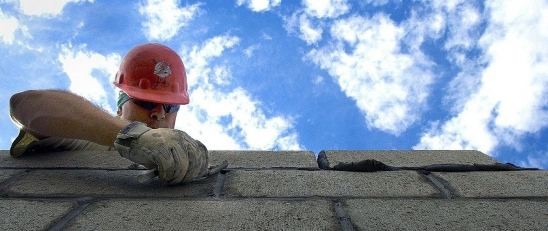 Pracownik budowlany w czerwonym kasku układa cegły na tle nieba z chmurami.