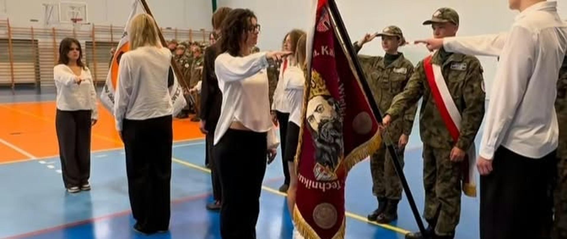 Uczniowie zwróceni twarzami do siebie z wyciągniętymi prawymi rękami przed siebie śrubują wobec pocztów sztandarowych dwóch szkół