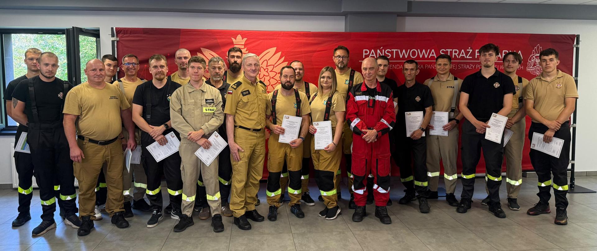 Grupowe zdjęcie uczestników szkolenia w umundurowaniu PSP, częściowo z wręczonymi zaświadczeniami, przed czerwonym banerem Państwowej Straży Pożarnej.
