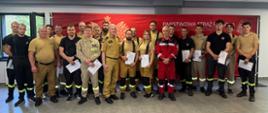 Grupowe zdjęcie uczestników szkolenia w umundurowaniu PSP, częściowo z wręczonymi zaświadczeniami, przed czerwonym banerem Państwowej Straży Pożarnej.
