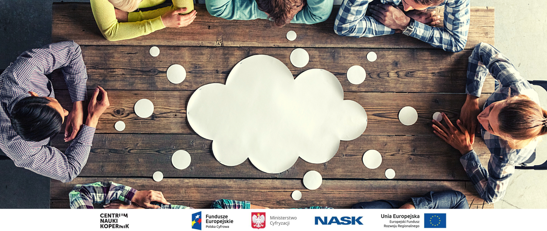 Kilka osób siedzących przy drewnianym stole (widok z góry). Na środku stoły biała chmura symbolizująca pomysły, myślenie, kreatywność.