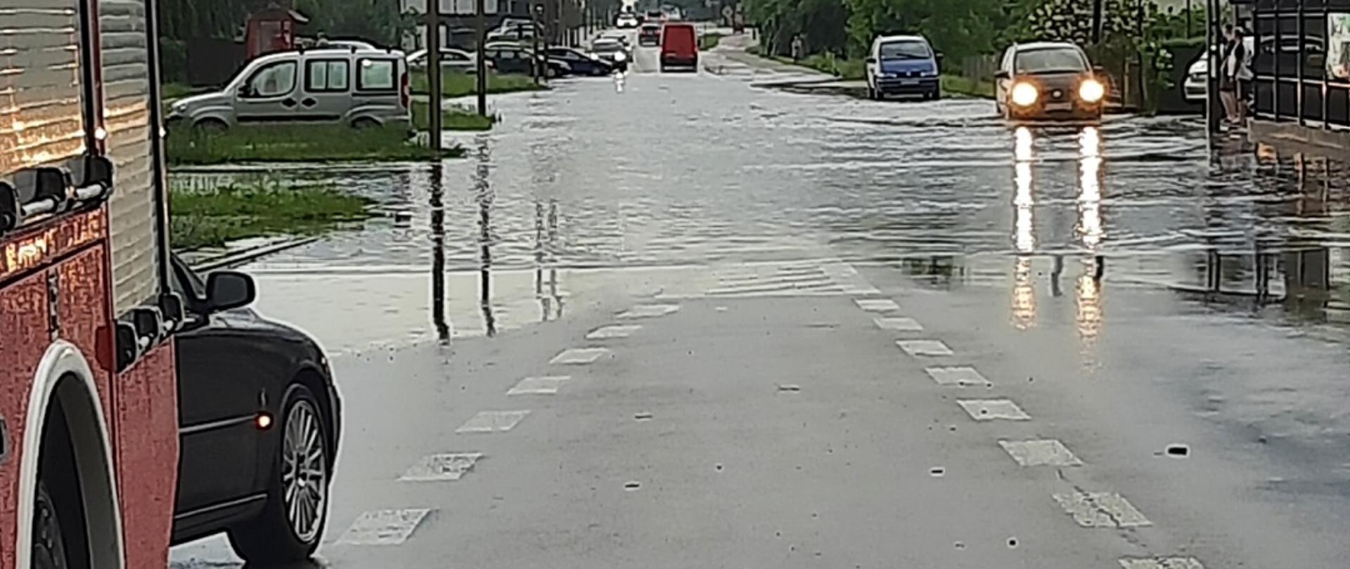 Zdjęcie obrazuje zalaną ulicę i przejeżdżające samochody