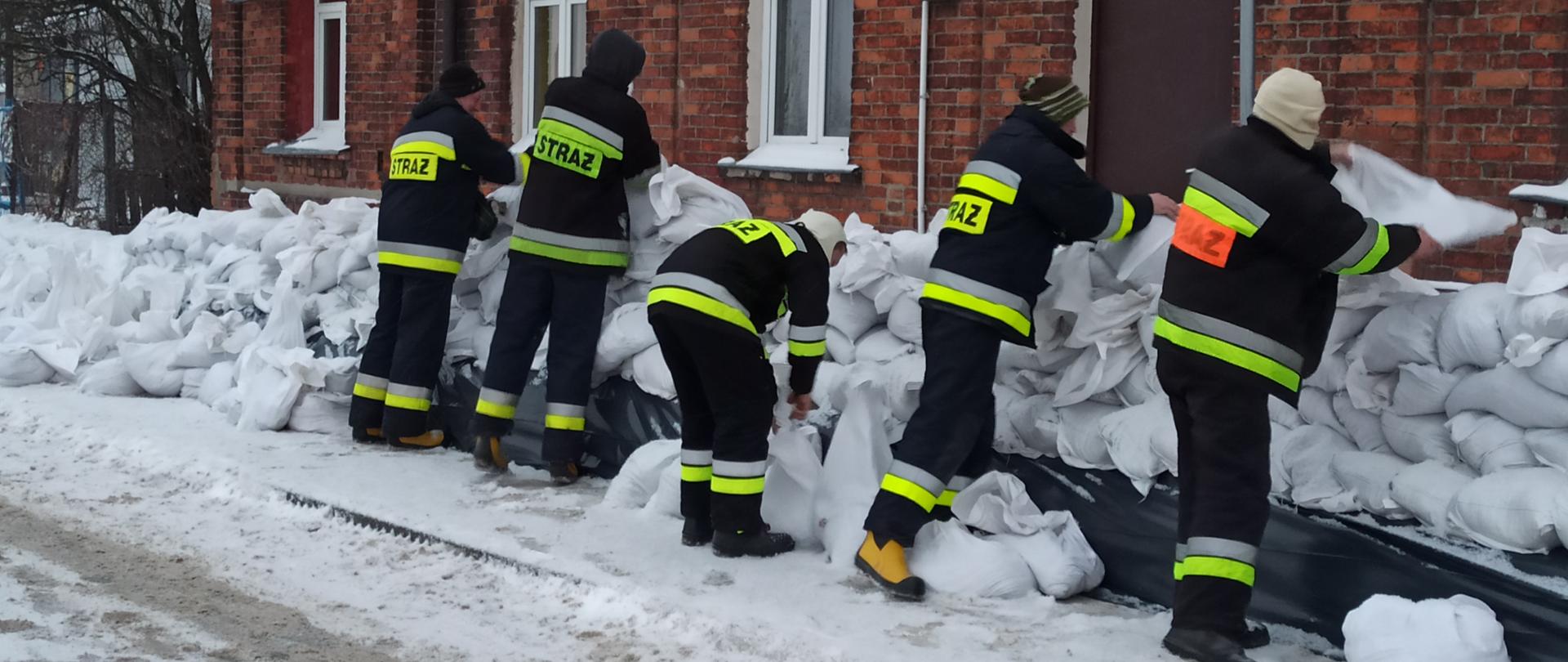 Na zdjęciu widać strażaków, którzy układają obwałowanie budynku z worków wypełnionych piaskiem