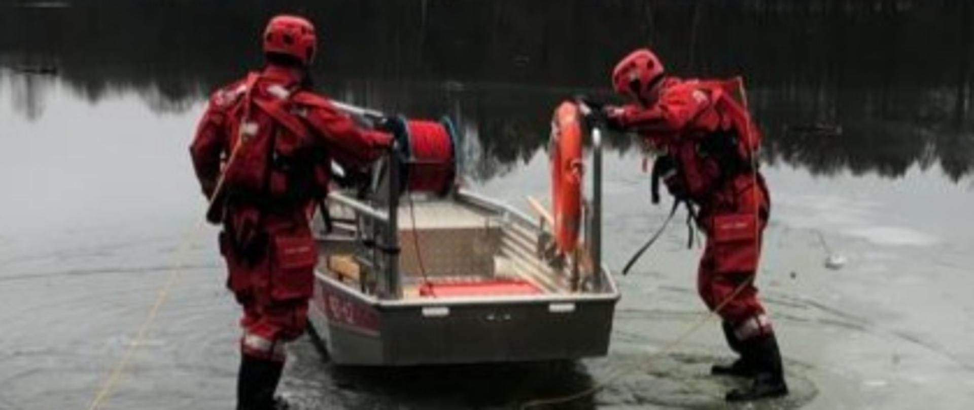 Na tafli zamarzniętego jeziora dwóch strażaków ubranych w specjalne suche skafandry do ratownictwa wodnego porusza się z aluminiową łodzią płaskodenną, która jest specjalnie wyposażona do przeprowadzania akcji na wodzie jak i na lodzie.
