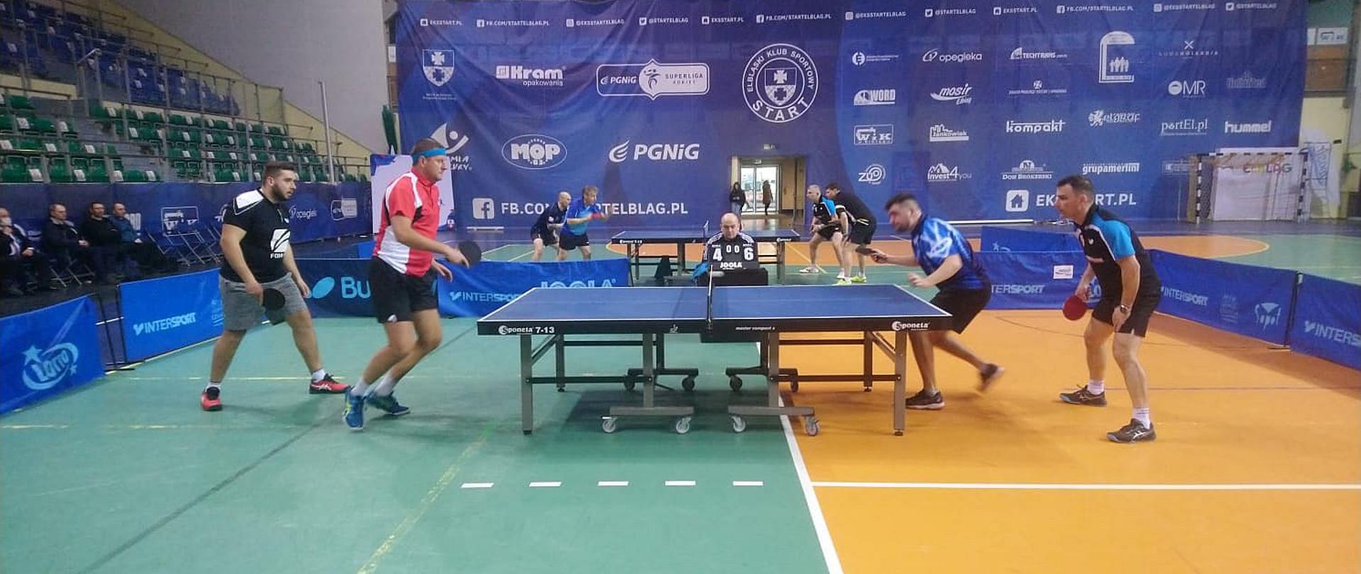 Rozgrywa meczu w tenisie stołowym rozgrywana w parach, po obu stronach stołu stoi po dwóch zawodników. W tle tocząca się rozgrywka przy innym stole.