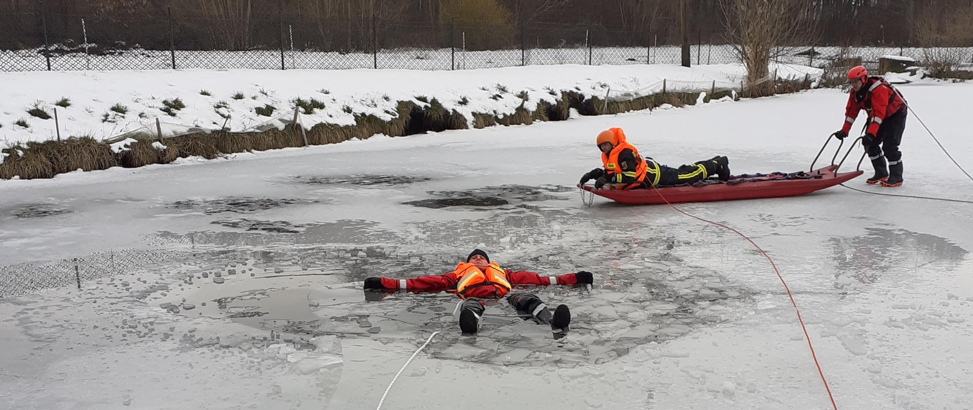 Na pierwszym planie widać osobę ubrana w kamizelkę ratunkową, pod którą załamał się lód. Osoba jest w wodzie i pływa na plecach. Za tą osobą widać dwóch strażaków ubranych w kamizelki ratunkowe, jeden z nich leży na saniach lodowych, drugi który je pcha.