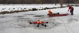 Na pierwszym planie widać osobę ubrana w kamizelkę ratunkową, pod którą załamał się lód. Osoba jest w wodzie i pływa na plecach. Za tą osobą widać dwóch strażaków ubranych w kamizelki ratunkowe, jeden z nich leży na saniach lodowych, drugi który je pcha.