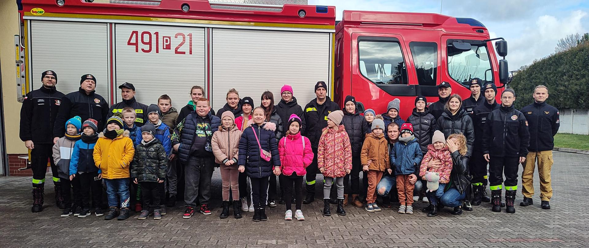 Zdjecie grupowe przedstawiające członków młodzieżowej drużyny pożarniczej w Kuklinowie oraz krotoszyńskich strażaków. Grupa dzieci wraz z opiekunami wizytowała krotoszyńską strażnicę poznając zawód strażaka. Zdjęcie wykonane na tle wozu pożarniczego