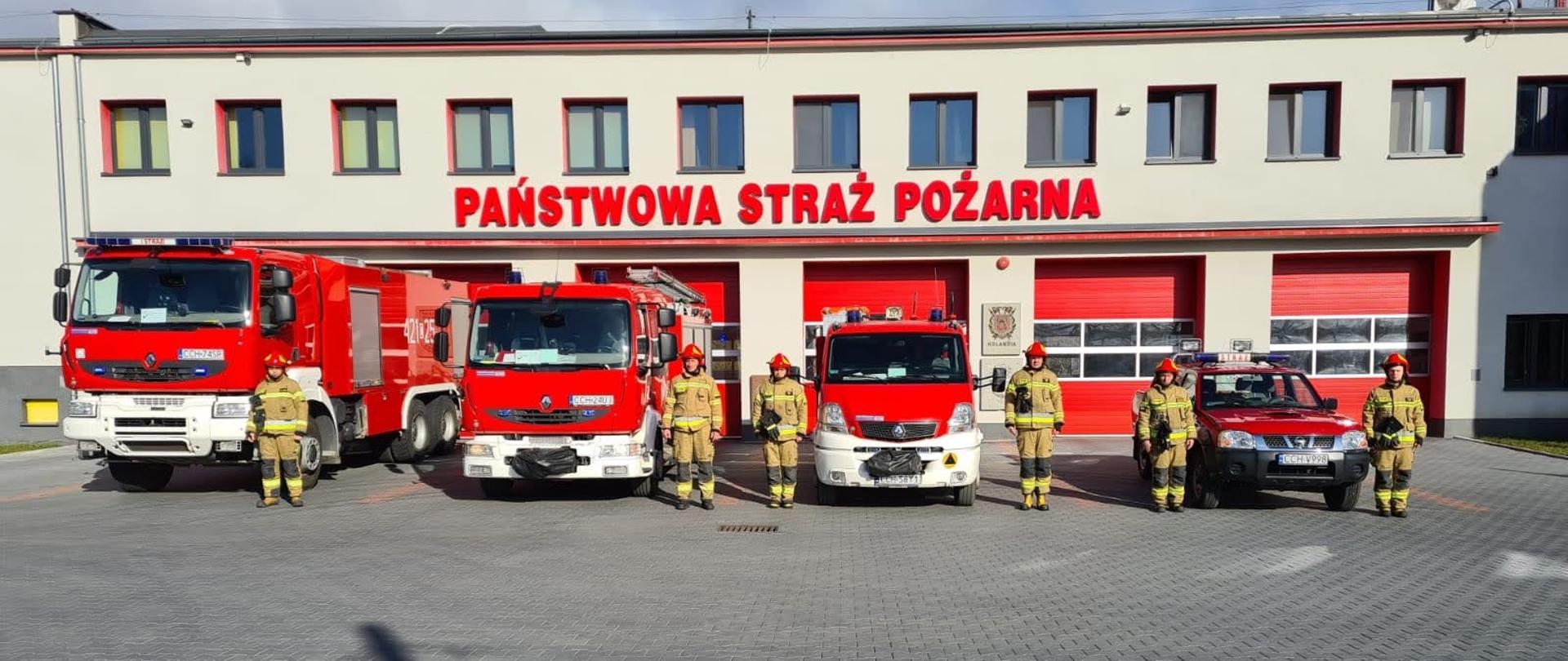 Na zdjęciu wykonanym przed budynkiem Komendy Powiatowej PSP w Chełmnie znajdują się strażacy stojący przy wozach bojowych w hołdzie ofiarom Katastrofy Smoleńskiej. Pojazdy mają uruchomione sygnały świetlne i dźwiękowe. 