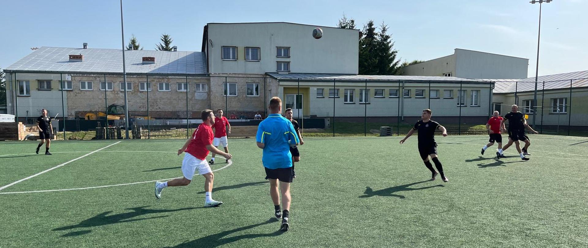 Na zdjęciu widać grupę mężczyzn grających w piłkę nożną na świeżym powietrzu. Znajdują się na boisku z syntetycznej trawy, otoczonym siatką, a w tle widać duży, dwupiętrowy budynek. Zawodnicy są ubrani w stroje sportowe, w różnych kolorach. Dwóch z nich, biegnie w kierunku piłki.