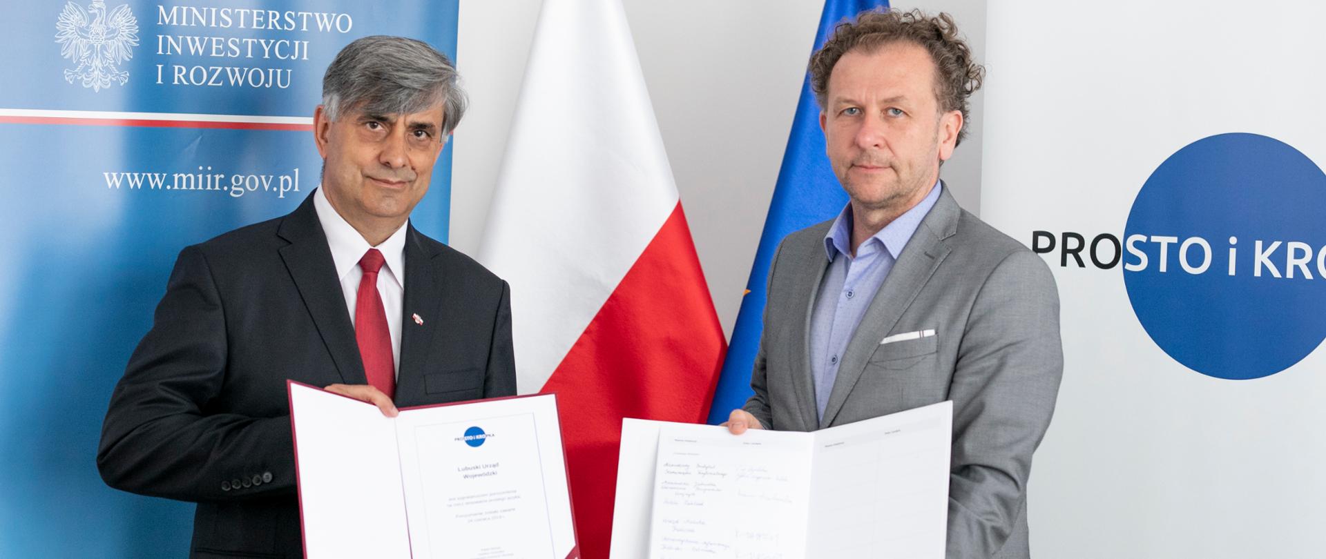 Dwóch mężczyzna stoi, w rękach trzymają podpisane umowy, za nimi znajduję się flaga Polska i Unii Europejskiej. Oraz rozkładane dwie ścianki z napisem "Prosto i Kropka" oraz "Ministerstwo Inwestycji i Rozwoju". 