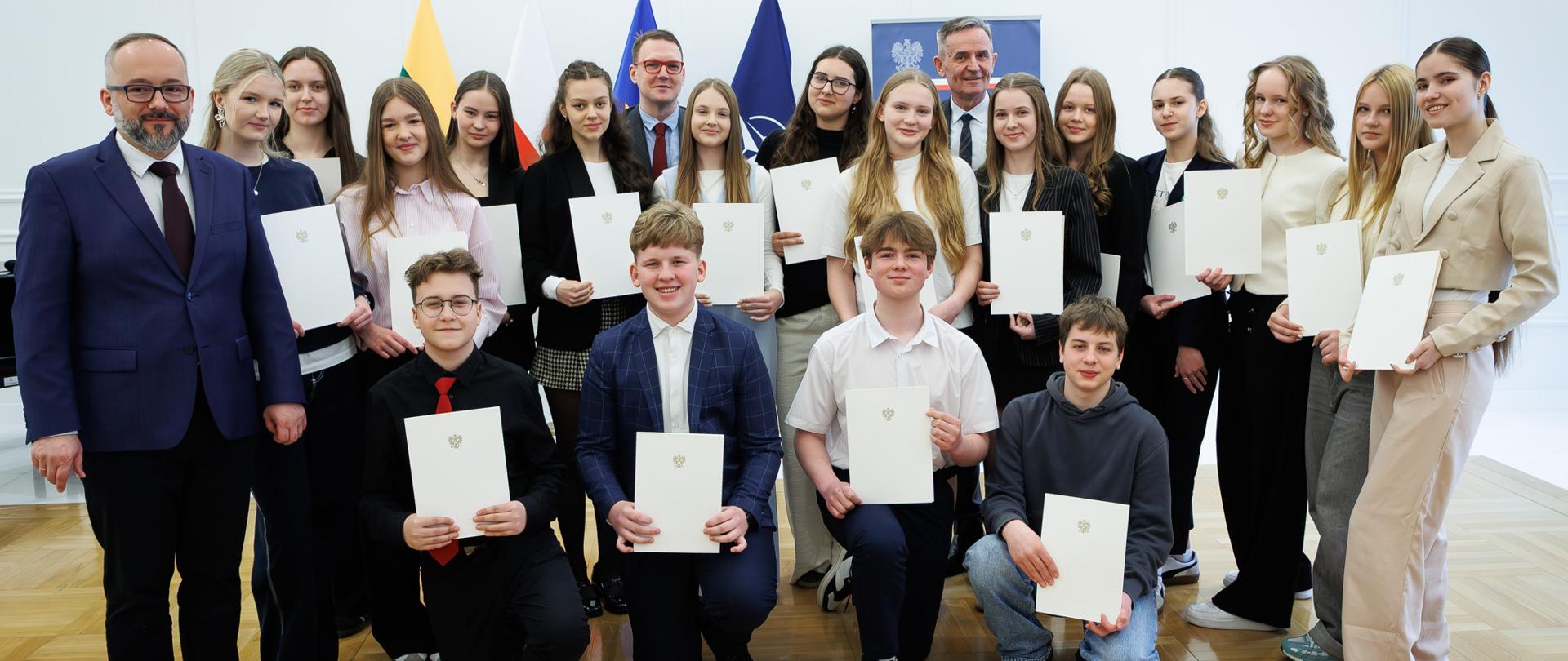 Nuotraukoje matoma didelė žmonių grupė -Jaunimo taryba prie Lenkijos Respublikos ambasados Vilniuje - sustojusi dviem eilėmis šviesioje salėje su medinėmis grindimis. Visi asmenys rankose laiko vienodus šviesius dokumentus su oficialiu herbu ant viršelio. Fone stovi kelios vėliavos.