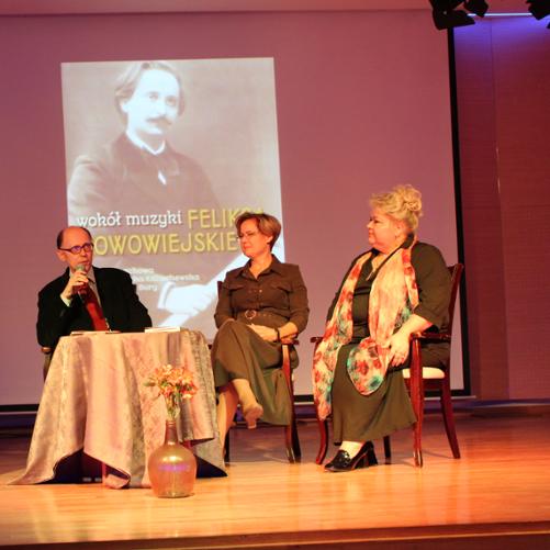 zdjęcie przedstawia organizatorów spotkania podczas wykładu na krzesłach przy stoliku na scenie sali koncertowej, w tle ekran multimedialny z portretem bohatera wykładu