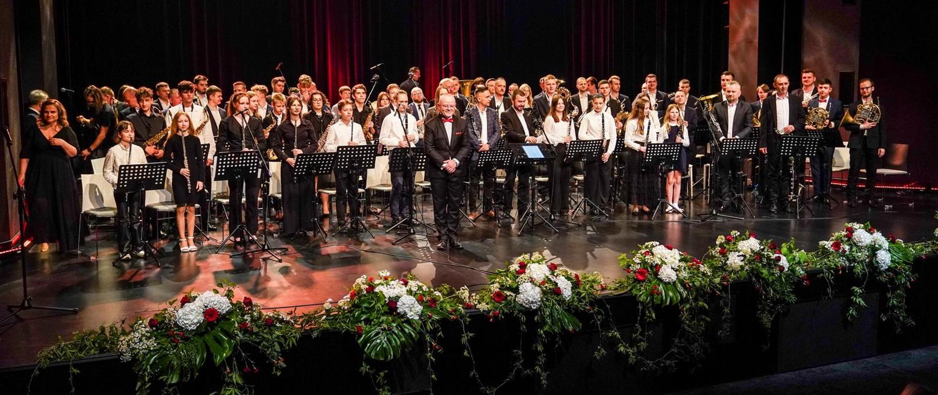 Orkiestra dęta stoi na scenie, dyrygent w pierwszym rzędzie, z przodu wiązanki kwiatów