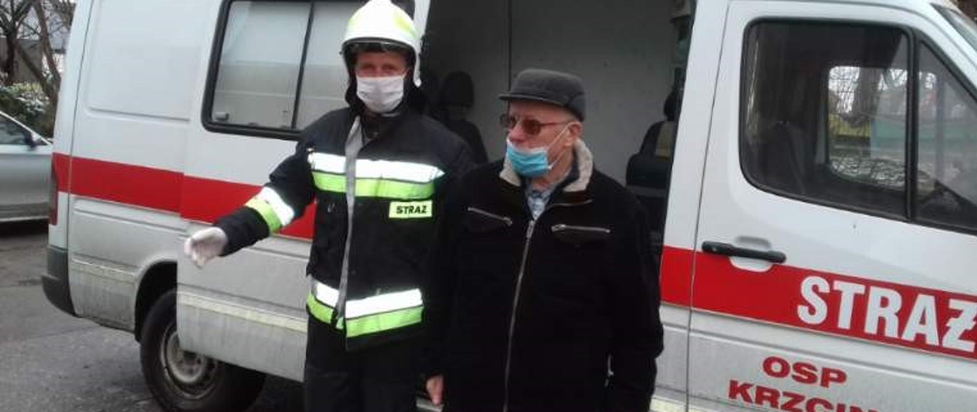 Zdjęcie przedstawia mężczyznę w maseczce i ubiorze zimowym w asyście strażaka ubranego w hełm strażacki, maseczkę białą i rękawiczki białe, po wyjściu z samochodu – busa strażackiego. Biały bus strażacki posiada poprzeczny czerwony pas. Przednie drzwi posiadają napis na czerwonym pasie biały napis „STRAŻ” natomiast niżej na białym tle czerwony napis OSP KRZCIN.