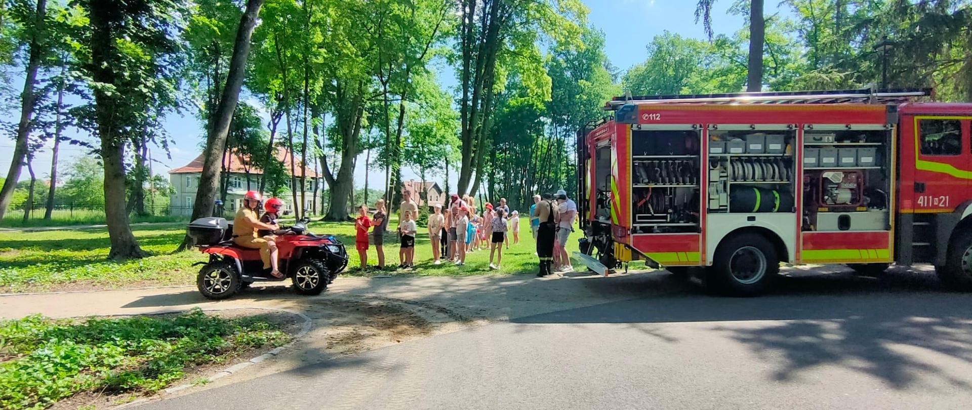 Park, zielone drzewa, słonecznie. Z prawej strony stoi samochód strażacki, otwarte skrytki. Z lewej pojazd kład strażacki. Przy pojazdach dzieci ze strażakami.