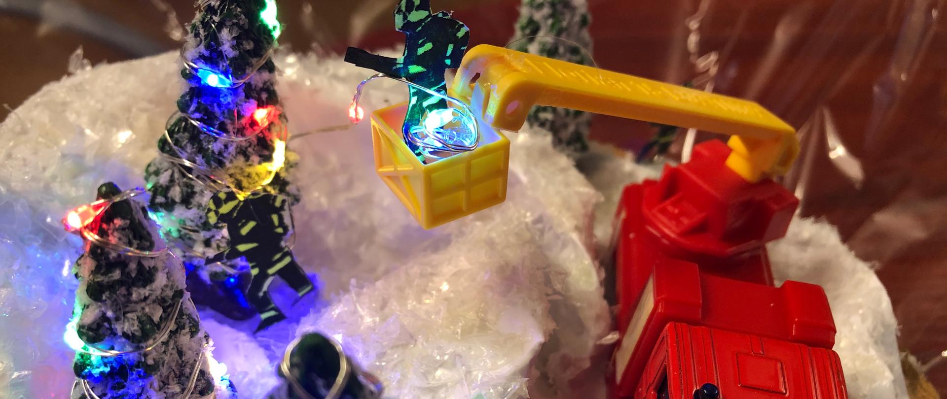 Stroik świąteczny na którym jest samochód drabina na niej strażak i trzy choinki oświetlone 