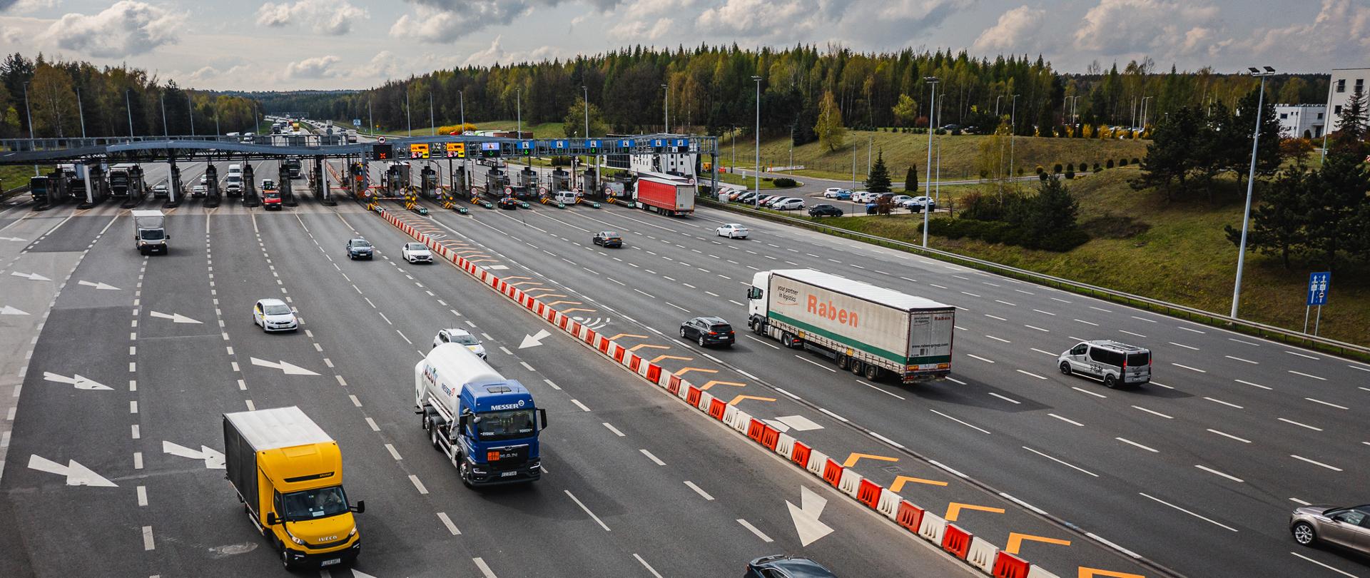 Widok z góry na wielopasmowy punkt poboru opłat na autostradzie A4. Przez bramki przejeżdżają samochody osobowe i ciężarowe, a na jezdni widoczne są liczne oznaczenia kierunkowe oraz separatory pasów ruchu. W tle znajduje się zalesiony teren i częściowo zachmurzone niebo. Na zdjęciu widoczna jest infrastruktura autostradowa związana z poborem opłat.
