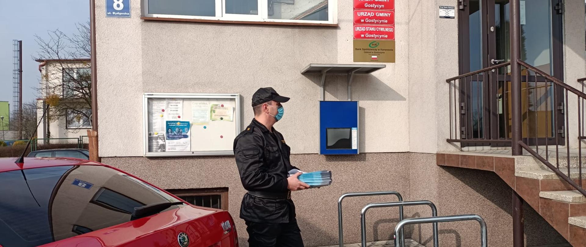 Funkcjonariusz trzymający pakiet ulotek przed siedzibą Urzędu Gminy Gostycyn. 