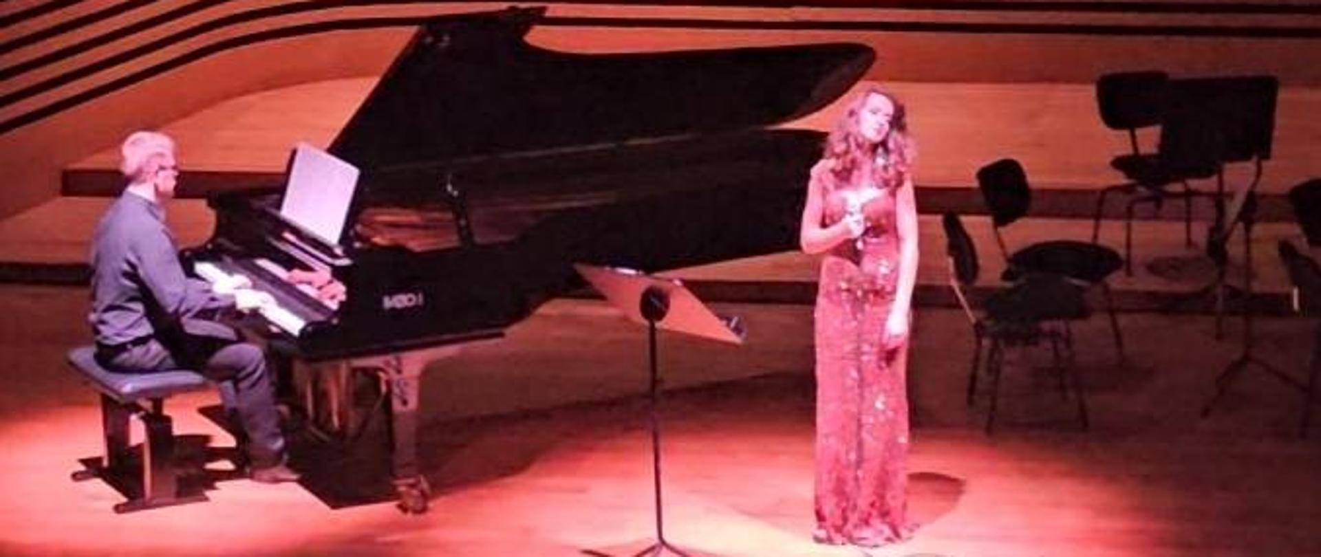 Kobieta śpiewa, mężczyzna akompaniuje jej na fortepianie.