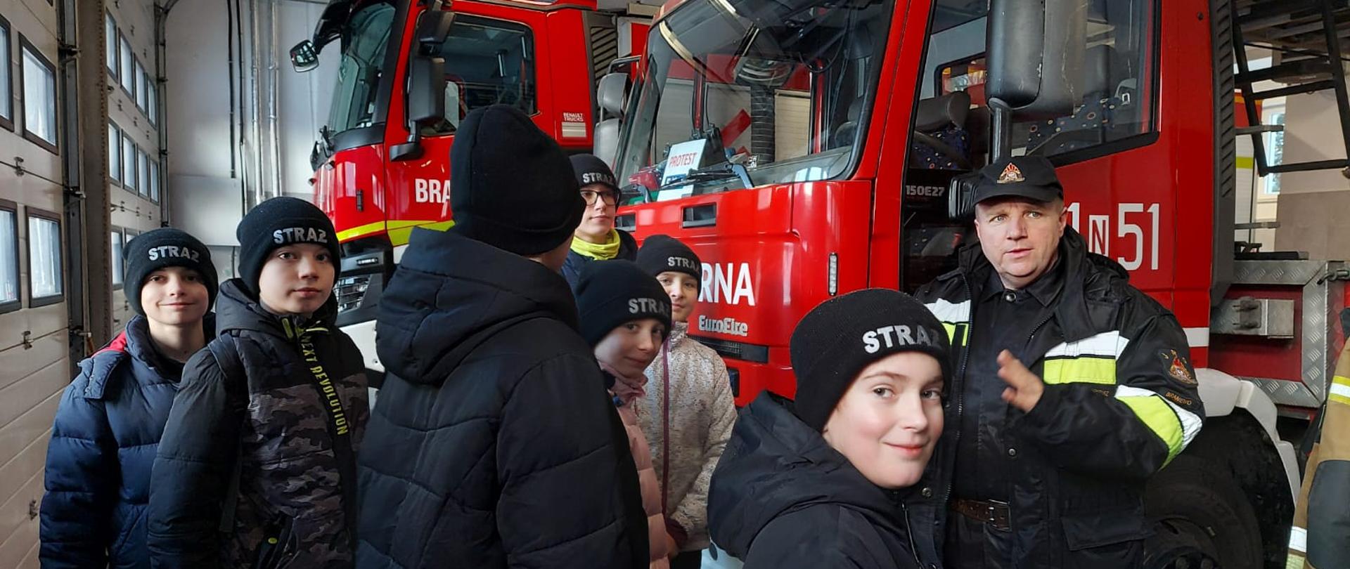 Zdjęcie zrobione w garażu bojowym. Na zdjęciu grupka dzieci i strażak. W tle czerwone samochody strażackie.