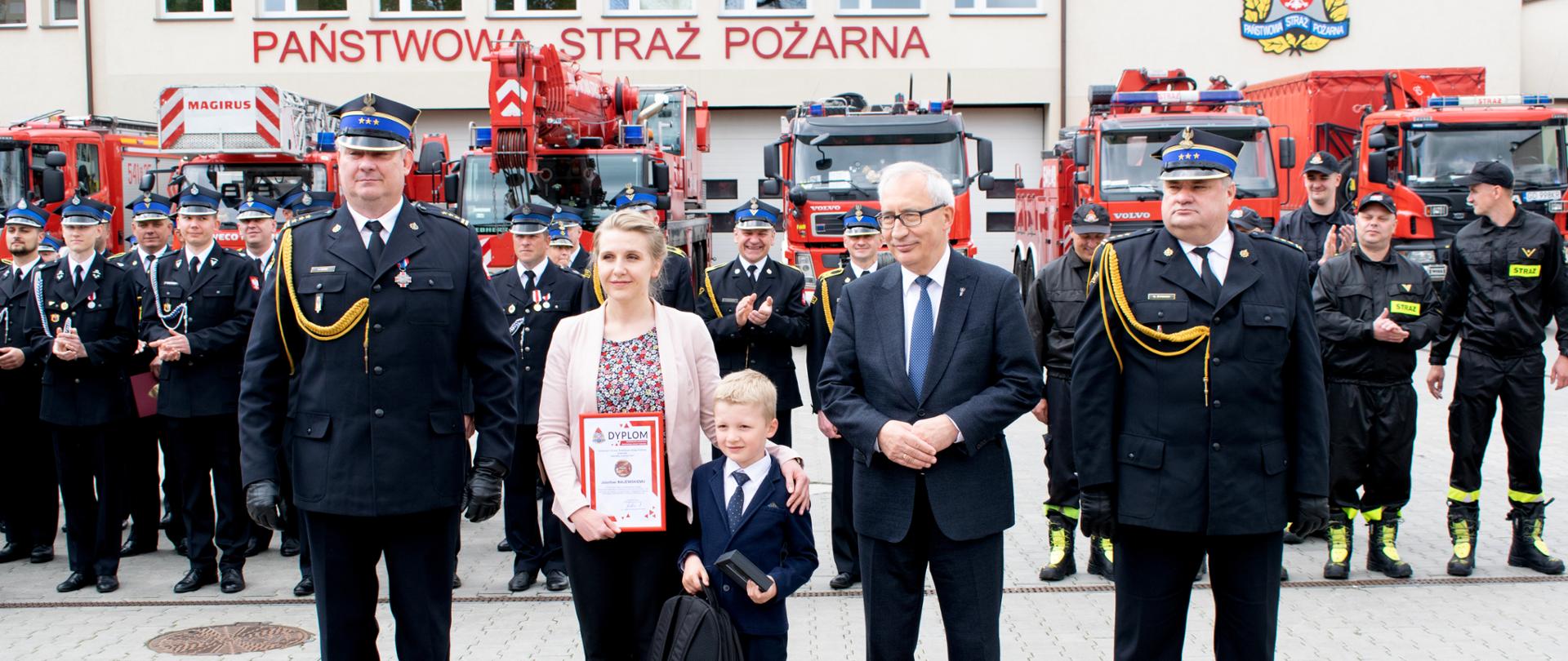 Plac KP PSP. Pierwszy plan - w szeregu stoją kolejno Komendant Powiatowy, mam odznaczonego dziecka, odznaczony Józio, Poseł na Sejm RP, Zastępca Komendanta Wojewódzkiego PSP. Drugi plan - pododdziały strażaków w umundurowaniu wyjściowym oklaskują odznaczonego. Trzeci plan - wozy strażackie ustawione przed garażami.
