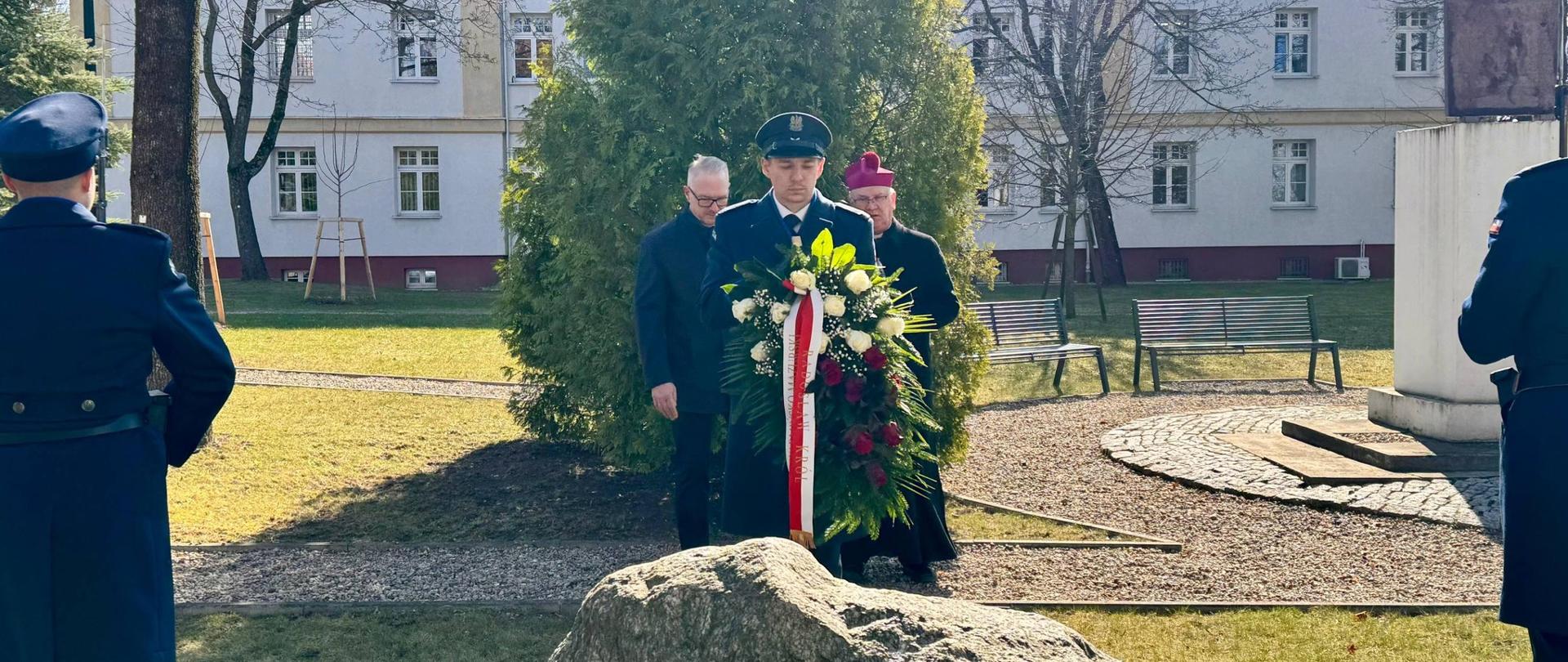 Uroczystości upamiętniające ofiary Zbrodni Katyńskiej: wojewoda i arcybiskup składają kwiaty, policjant trzyma wiązankę biało-czerwonych kwiatów.