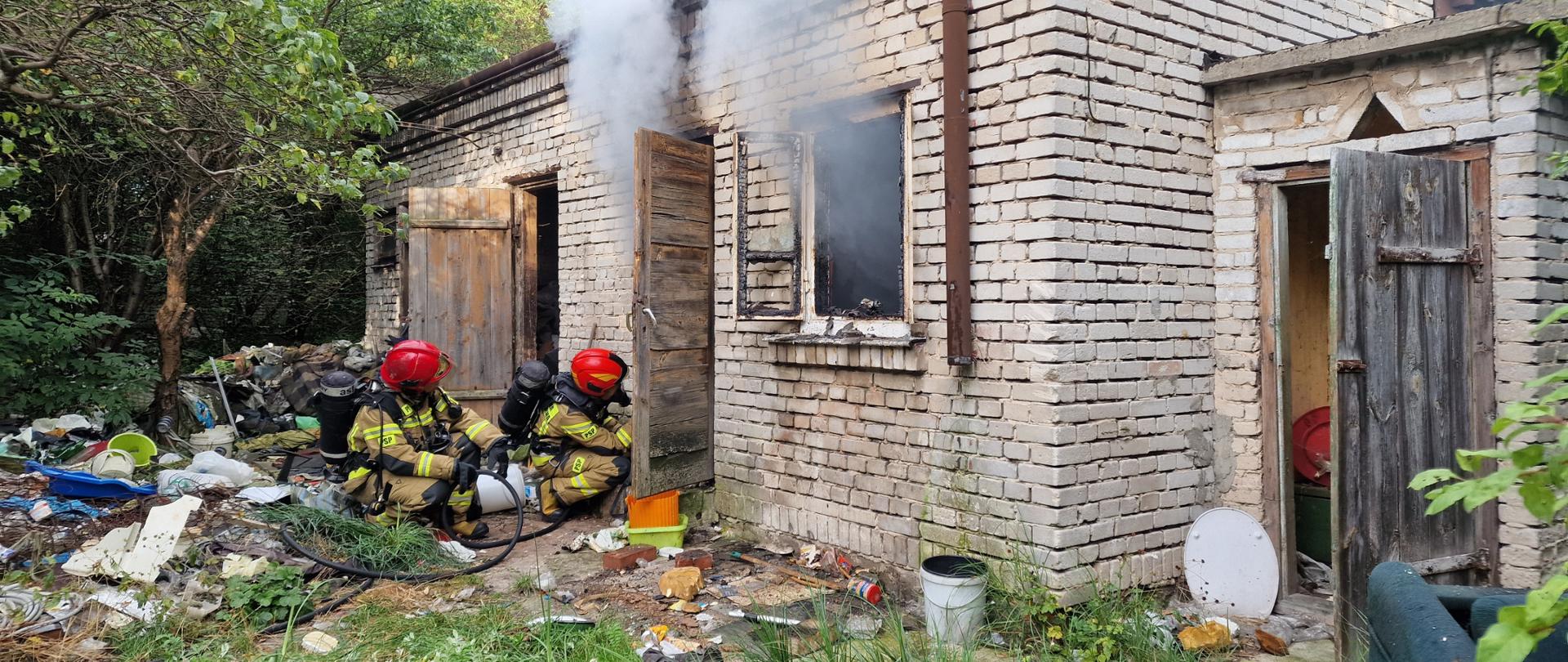 Na pierwszym planie dwóch strażaków klęczy przed wejściem do budynku i gasi pożar 
